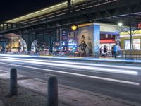 an intersection with many shops in the distance at night time, and a bus passing under an elevated overpass