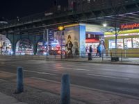 an intersection with many shops in the distance at night time, and a bus passing under an elevated overpass