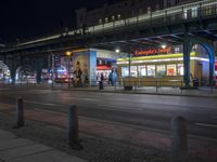 an intersection with many shops in the distance at night time, and a bus passing under an elevated overpass