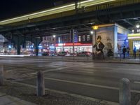 an intersection with many shops in the distance at night time, and a bus passing under an elevated overpass