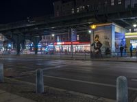 an intersection with many shops in the distance at night time, and a bus passing under an elevated overpass