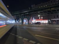 an intersection with many shops in the distance at night time, and a bus passing under an elevated overpass