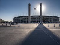 the sun is shining over a large building at an olympics venue with people standing on the sidelines and spectators in the distance