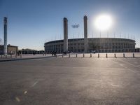 the sun is shining over a large building at an olympics venue with people standing on the sidelines and spectators in the distance