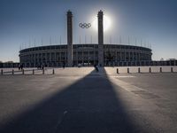 the sun is shining over a large building at an olympics venue with people standing on the sidelines and spectators in the distance