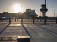 the sun is shining on a city square area with benches and gates near an airplane control tower