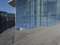 a large blue glass building next to water and buildings with blue windows and grey concrete flooring