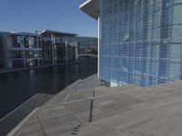 a large blue glass building next to water and buildings with blue windows and grey concrete flooring