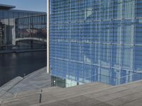 a large blue glass building next to water and buildings with blue windows and grey concrete flooring