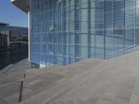 a large blue glass building next to water and buildings with blue windows and grey concrete flooring
