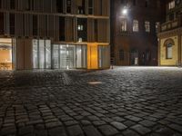 empty, cobblestone courtyard with tall buildings at night in the background, with lighting on