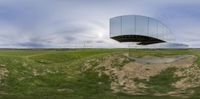 a reflection of a curved building on a green field under a blue cloudy sky and in the distance it's a field that has grass