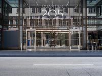 glass facade entrance of the hotel with silver lettering and people walking through it, near street corner, and two tall buildings