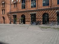 a very wide and empty brick sidewalk next to many bicycles in the background, a building with multiple windows