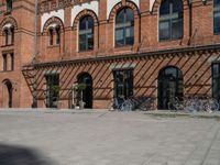 a very wide and empty brick sidewalk next to many bicycles in the background, a building with multiple windows