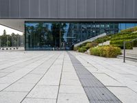 an empty stone walkway in front of a building with lots of windows and stairs with grass on the bottom