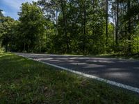 a asphalt road in front of wooded trees and blue sky with fluffy white clouds above