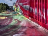 a graffiti painted walkway near green trees and road light poles next to a building with red and blue walls