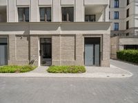 a building has plants in the front of it and bushes on the sidewalk with doors