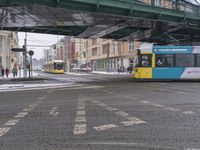 bus driving under an overpass in the snow by some buildings and people standing on a sidewalk