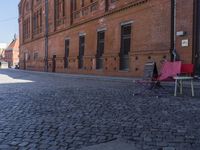 two pink chairs are sitting next to an empty brick walkway, with buildings in the background