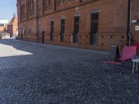 two pink chairs are sitting next to an empty brick walkway, with buildings in the background