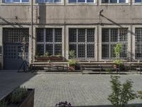 several benches in front of a concrete building with open window panes and potted plants