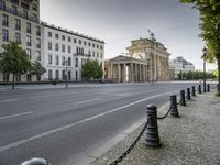 a city road with an old building in the background behind it, on the right side is a chain fence and a black post