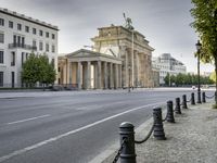 a city road with an old building in the background behind it, on the right side is a chain fence and a black post