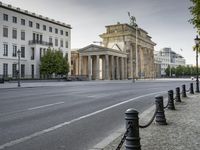 a city road with an old building in the background behind it, on the right side is a chain fence and a black post