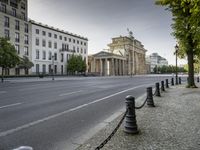 a city road with an old building in the background behind it, on the right side is a chain fence and a black post