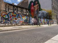 a wall with different colorful artwork on it in a city with buildings around and the street has a bike path going under it