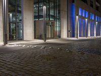 a brick paved sidewalk near a glass building at night, with street lights on and empty road, a light - up fire hydrant, blue in front