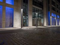 a brick paved sidewalk near a glass building at night, with street lights on and empty road, a light - up fire hydrant, blue in front