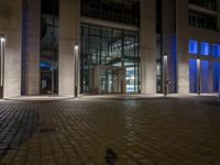 a brick paved sidewalk near a glass building at night, with street lights on and empty road, a light - up fire hydrant, blue in front