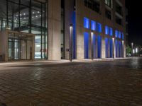 a brick paved sidewalk near a glass building at night, with street lights on and empty road, a light - up fire hydrant, blue in front