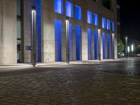 a brick paved sidewalk near a glass building at night, with street lights on and empty road, a light - up fire hydrant, blue in front