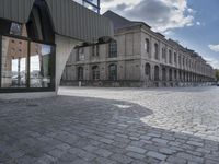 a cobble - stone sidewalk near large buildings, two people walking by it, on a cloudy day