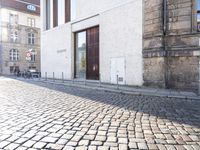 a woman riding a bike down a cobblestone street between buildings and sidewalk area