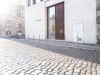 a woman riding a bike down a cobblestone street between buildings and sidewalk area