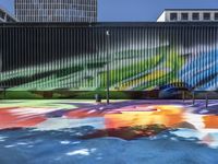 a colorful painted mural next to tall buildings and a bench in front of it at a sidewalk