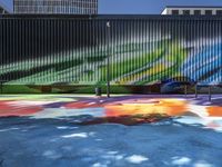 a colorful painted mural next to tall buildings and a bench in front of it at a sidewalk