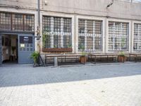 a row of benches sitting on a cobble stone street next to a building with windows and lots of plants