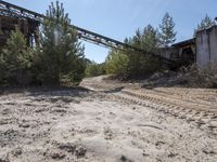a dirt road with tracks leading to an abandoned structure on a sunny day out with some trees and bushes in the background