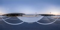 skateboarders are doing tricks on an empty asphalt surface at dusk with a circular ramp behind them