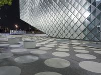 people sitting on white cement benches in front of an art museum, with lit up glass windows behind them