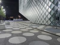 people sitting on white cement benches in front of an art museum, with lit up glass windows behind them