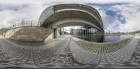 a fisheye photograph of a city under a bridge with water running through it and clouds in the sky