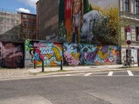 a wall with different colorful artwork on it in a city with buildings around and the street has a bike path going under it