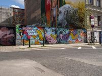 a wall with different colorful artwork on it in a city with buildings around and the street has a bike path going under it